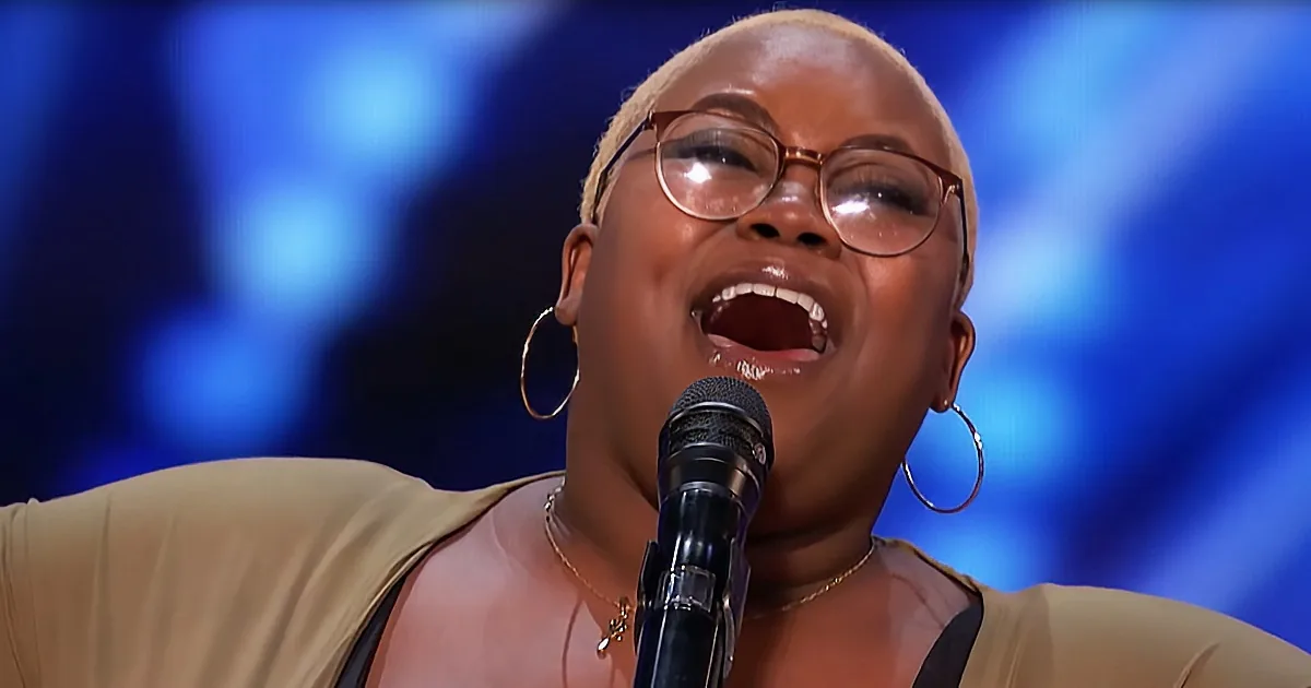 A woman with glasses and hoop earrings sings into a microphone, wearing a beige top.