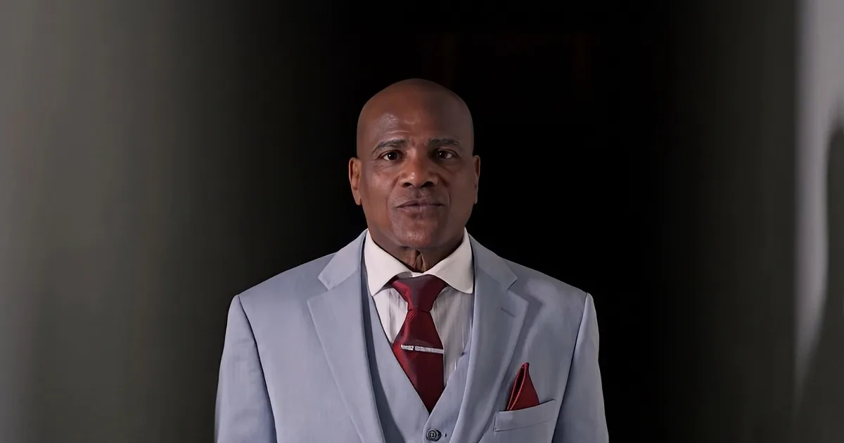 A man in a light gray suit with a red tie and pocket square stands against a dark background.