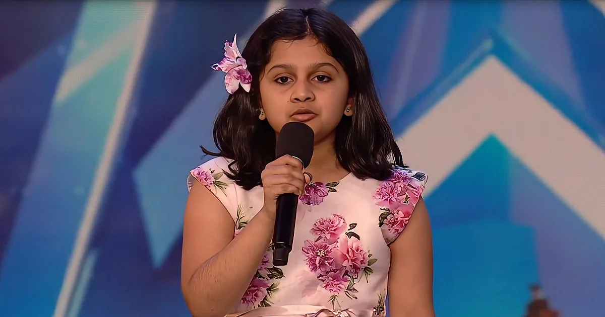 A young girl in a floral dress holds a microphone, standing on a stage with a blue geometric background.