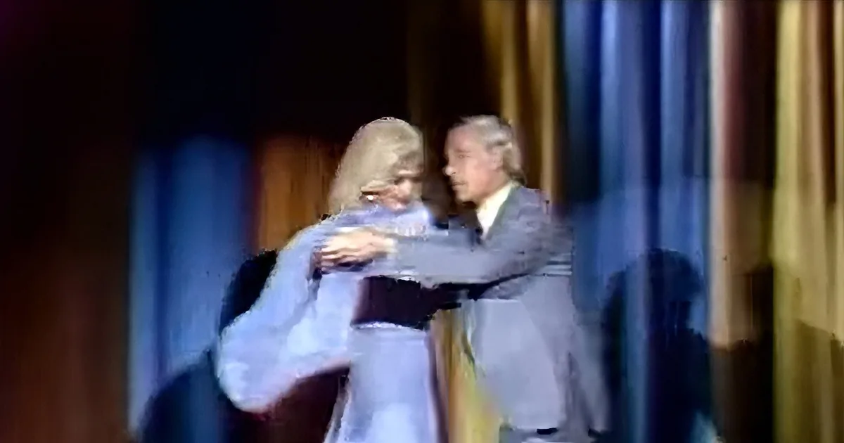 A man in a suit hugs a woman in a white dress on stage with a colorful backdrop.