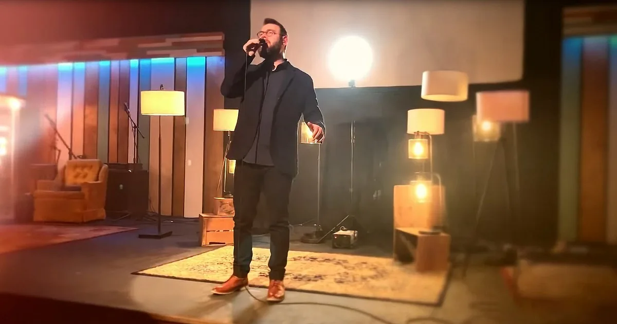 A man in a suit sings into a microphone on a stage with warm lighting and lamps.