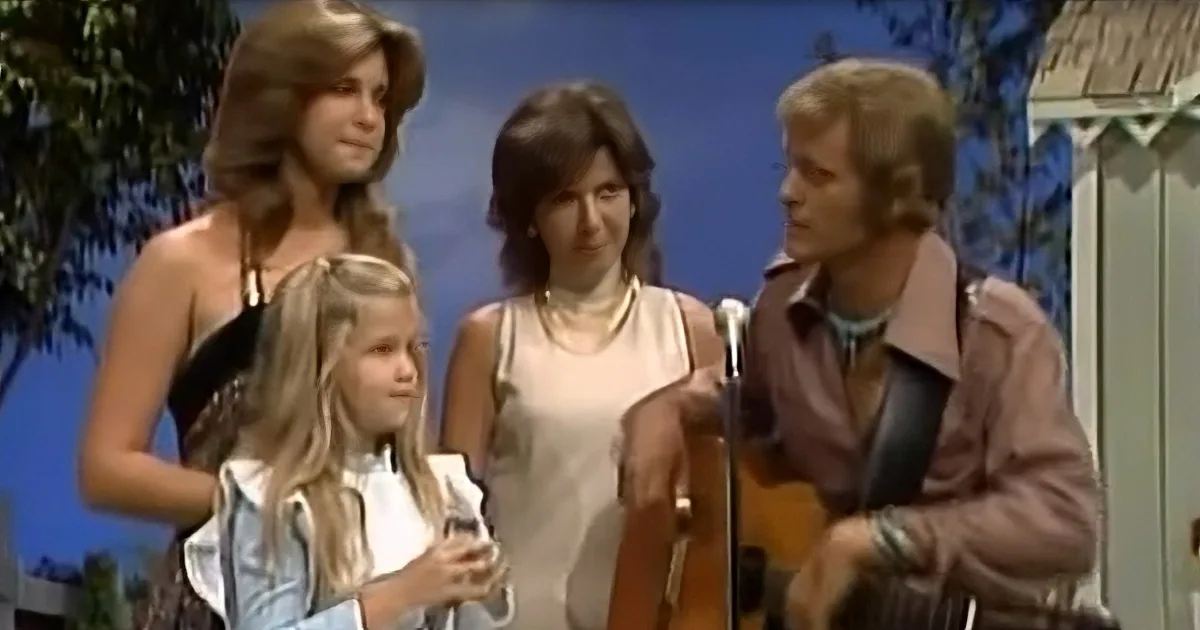A man plays guitar while a woman and two children watch, set against a blue sky backdrop.