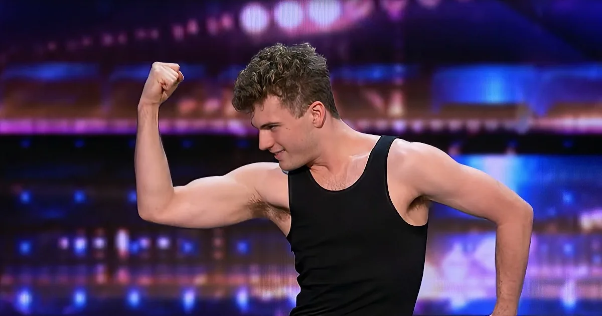 A man in a black tank top flexes his arm on a stage with colorful lights.