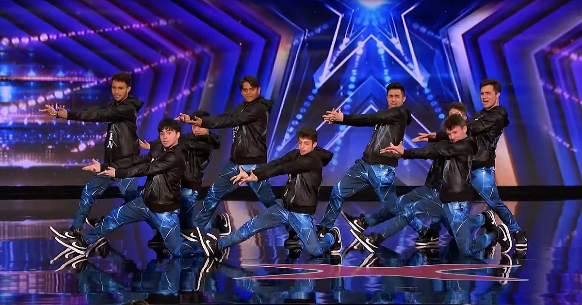 A group of dancers in black jackets and blue jeans perform a synchronized routine on a stage with a star-shaped backdrop.
