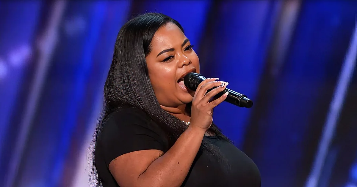A woman with long dark hair sings passionately into a microphone on stage.