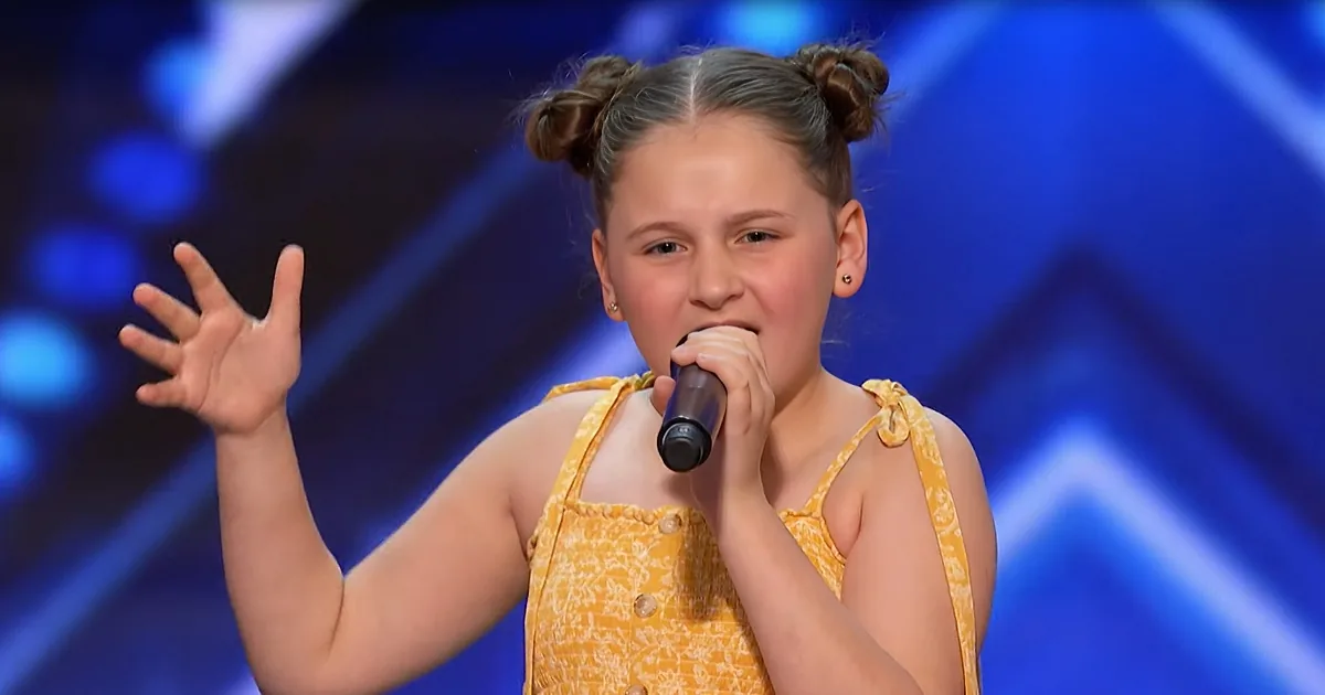 A young girl in a yellow dress sings into a microphone, her hand raised, against a blue stage backdrop.
