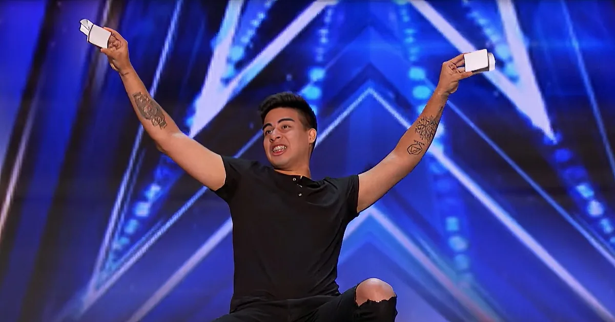 A person with tattoos raises their arms in celebration on a stage with blue lighting.