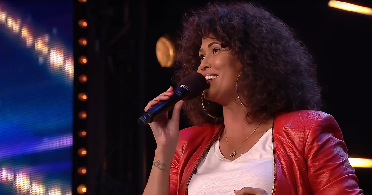 A woman with curly hair, wearing a red jacket, smiles while holding a microphone on stage.