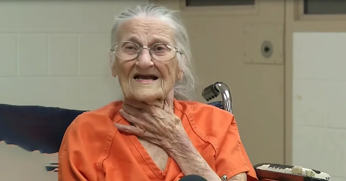 An elderly woman in an orange shirt sits in a wheelchair, touching her neck, with a plain wall and door in the background.