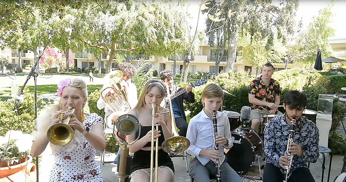 A group of musicians, including trumpet, trombone, clarinet, and drums, perform outdoors in a sunny garden setting.