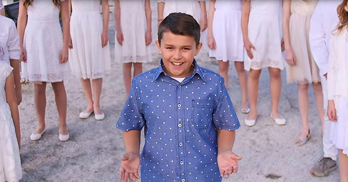 A boy in a blue polka-dot shirt smiles in front of a group of women in white dresses.