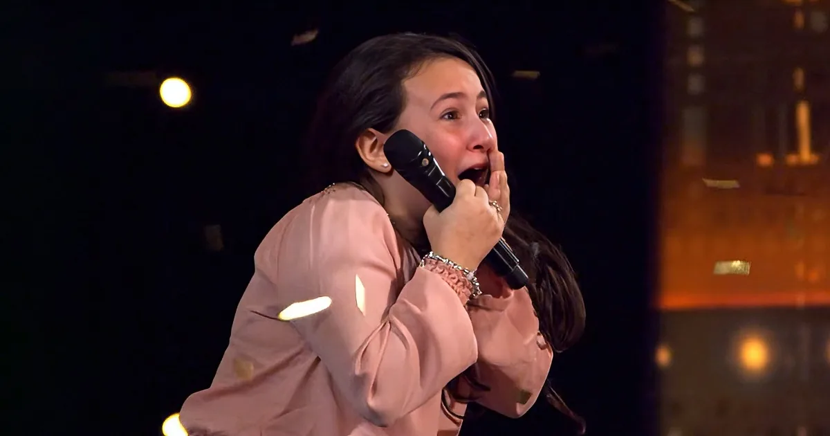 A woman in a pink outfit holds a microphone, her hand near her mouth, possibly singing or speaking.