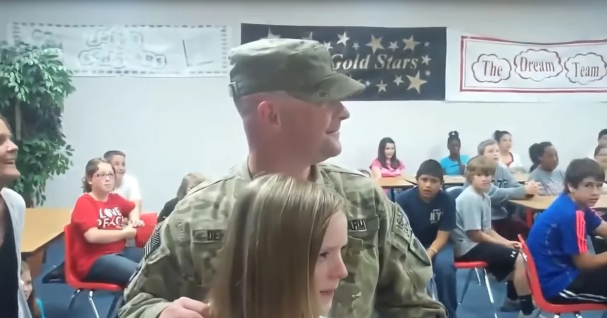 A soldier in uniform stands in front of a classroom of seated students, with banners in the background.