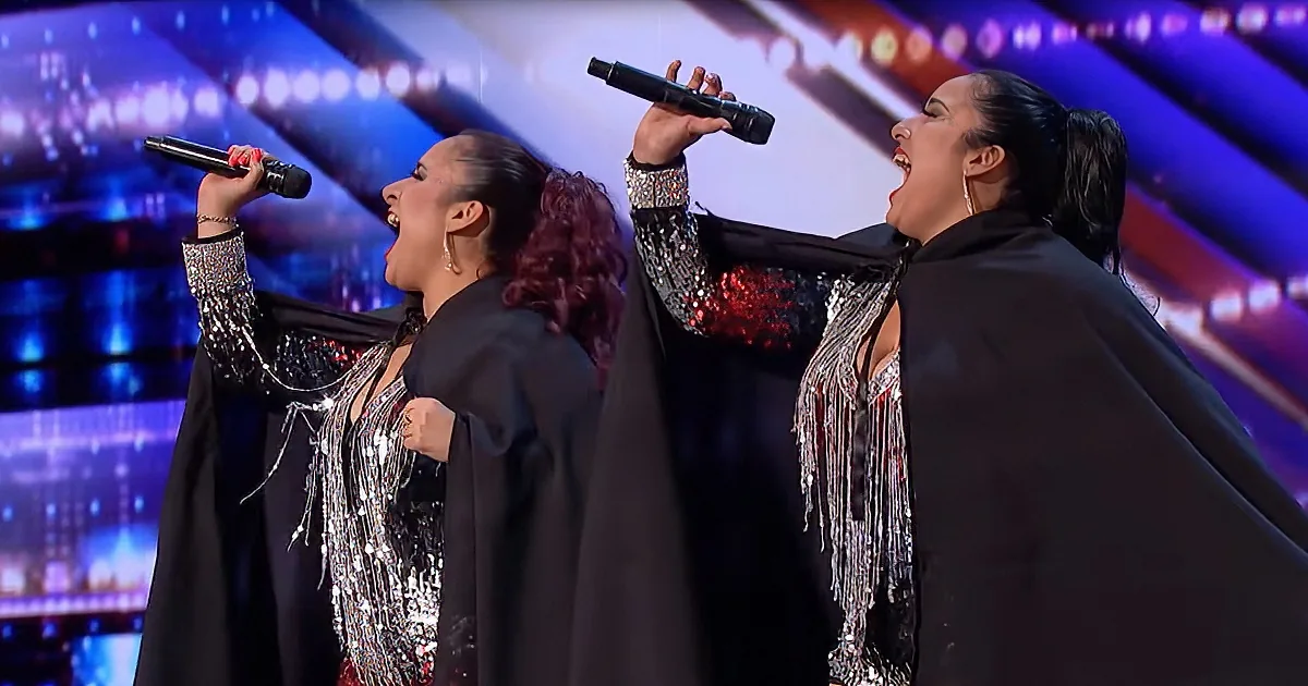 Two women in black sequined outfits sing passionately on stage with vibrant lights behind them.