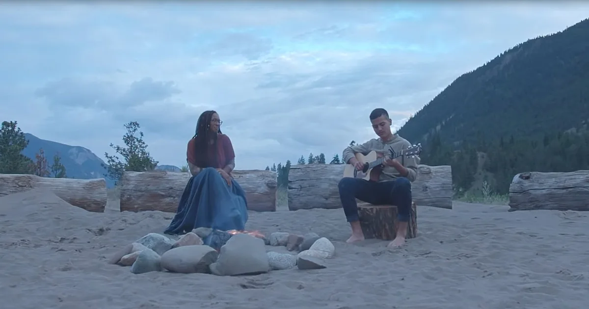 A man plays guitar while a woman sits nearby by a campfire on a sandy beach with mountains in the background.