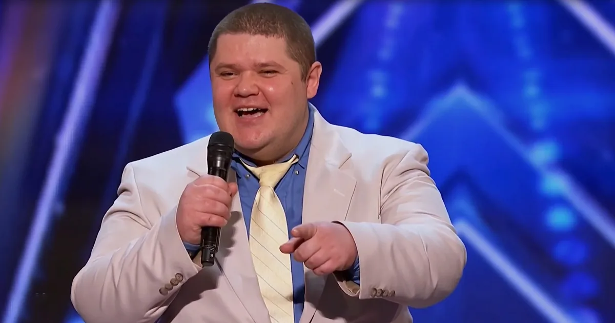 A man in a white suit and yellow tie holds a microphone, pointing, against a blue stage backdrop.
