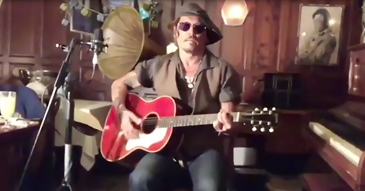 A man in a hat and sunglasses plays a red guitar in a cozy, dimly lit room.
