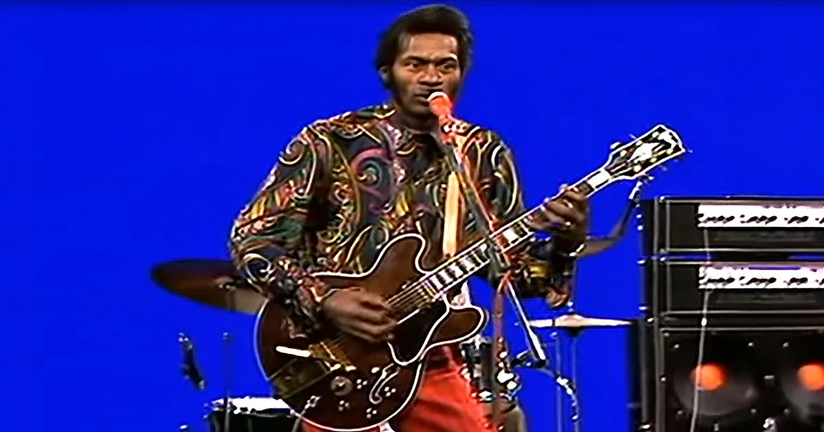A musician in a colorful shirt plays a guitar and sings into a microphone on stage.