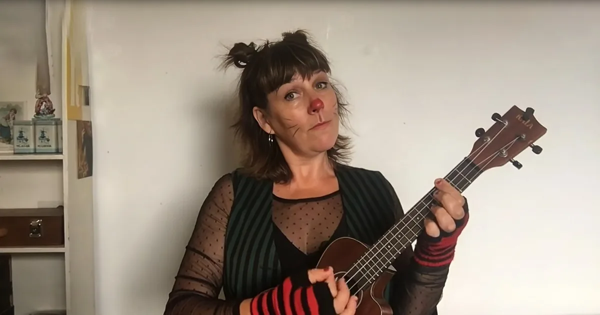A woman with a red nose and cat whiskers holds a ukulele, wearing striped sleeves and a green striped top.