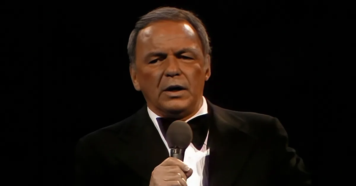 A man in a suit and bow tie holds a microphone, speaking against a dark background.