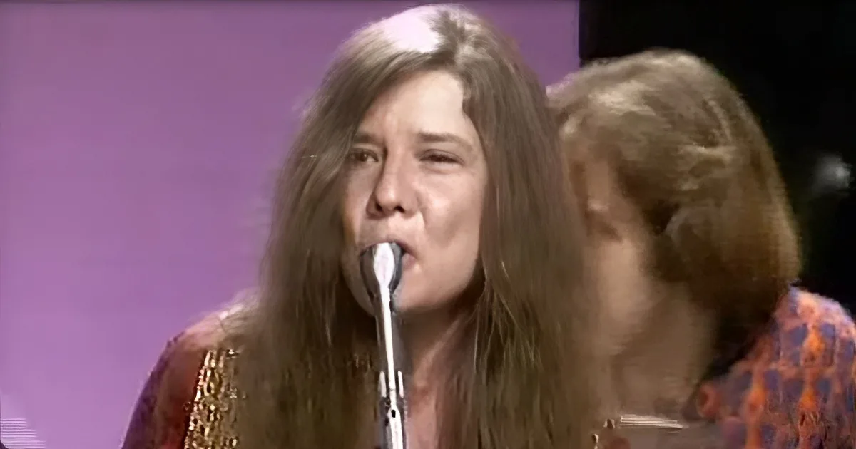 A woman with long hair sings into a microphone, wearing a patterned top, with another person partially visible beside her.