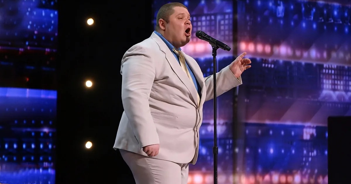 A man in a white suit sings passionately on stage, illuminated by stage lights.