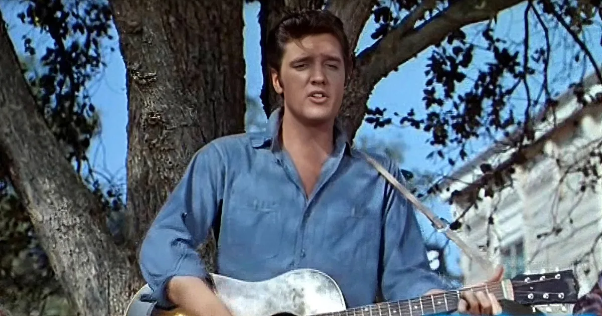 Elvis Presley in a blue shirt plays a guitar outdoors, surrounded by trees and a building.