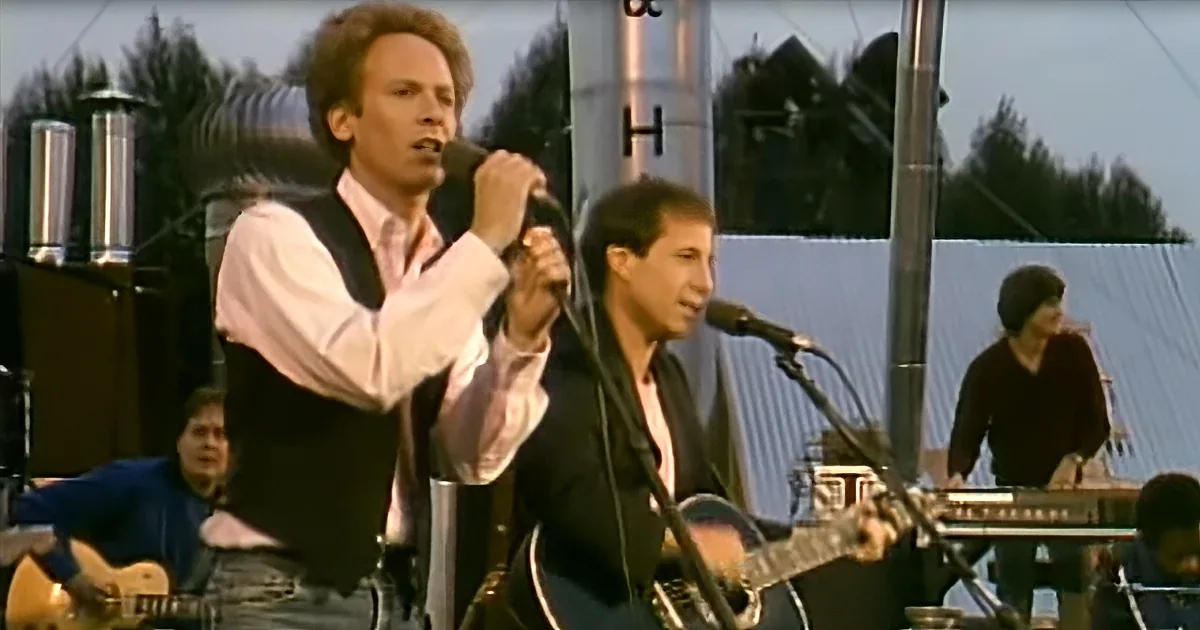 A man in a white shirt and black vest sings into a microphone on stage with musicians behind him.