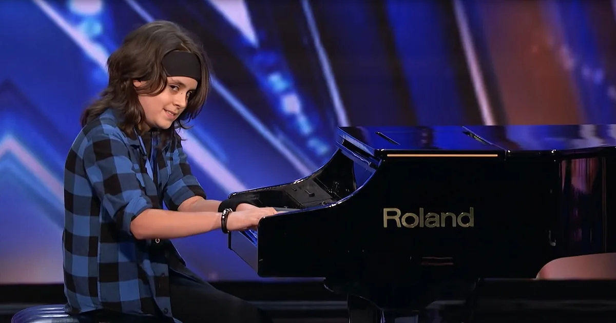 A young person in a plaid shirt plays a black Roland piano on a stage with blue lighting.