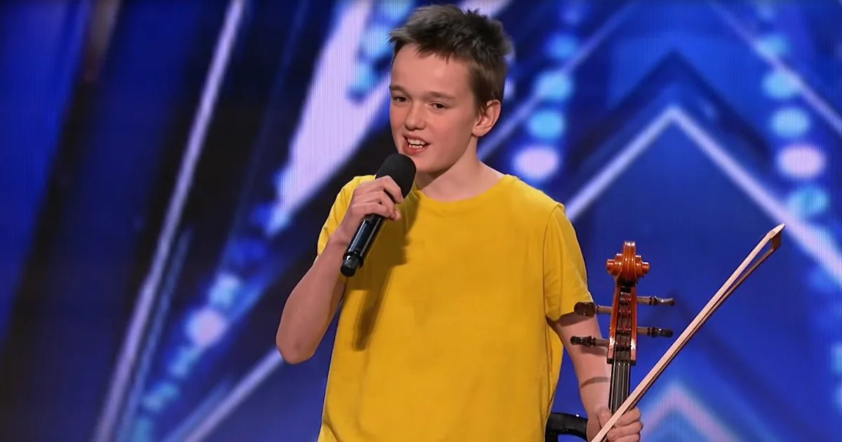 A young boy in a yellow shirt holds a microphone, smiling, with a cello in the background.