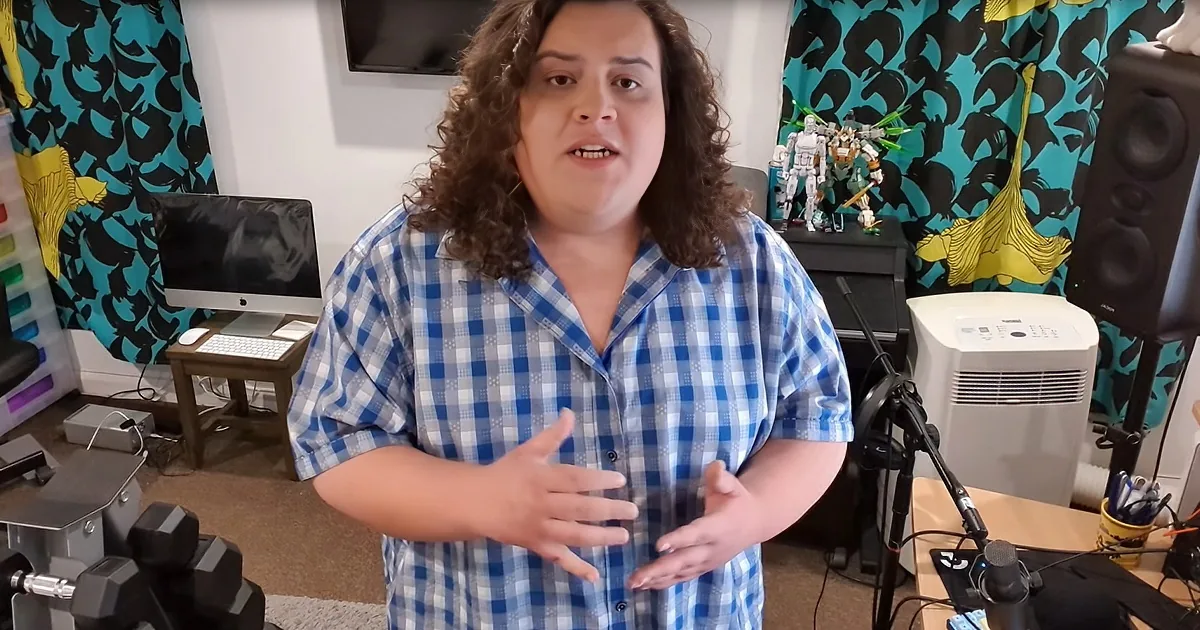 A person in a blue checkered shirt gestures with hands, surrounded by tech, decor, and a monitor in the background.
