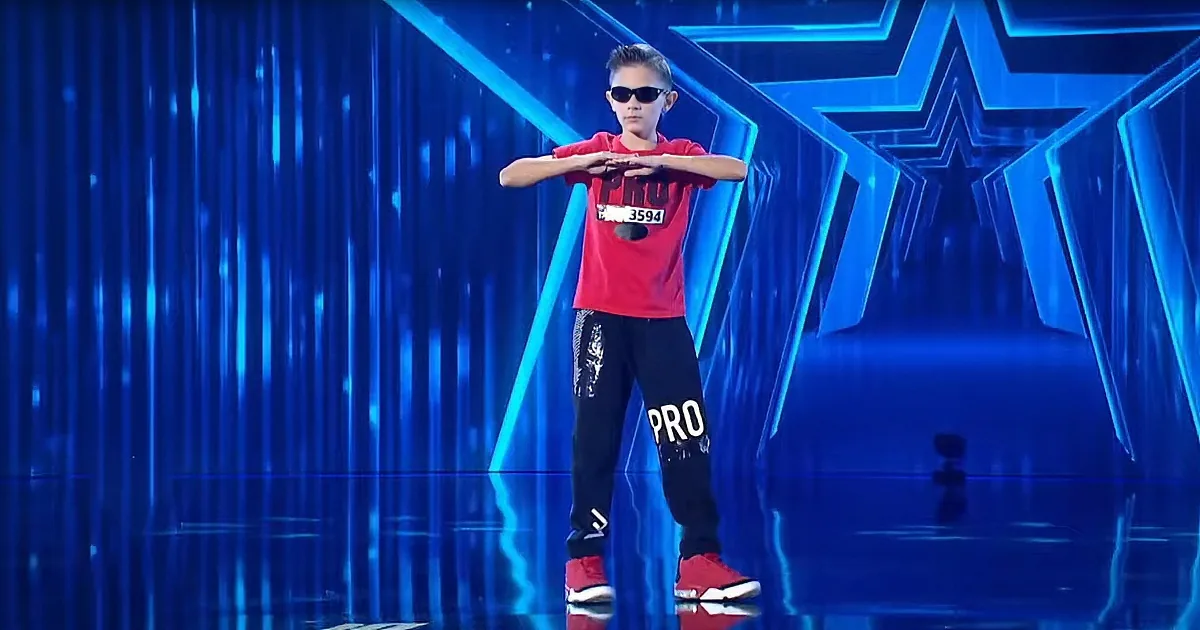 A young boy in a red shirt and black pants stands on stage with his arms crossed, wearing sunglasses and red shoes.