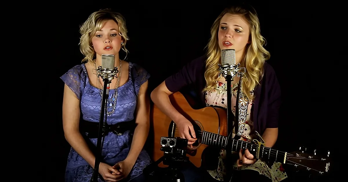 Two women sing into microphones, one plays guitar, both are dressed casually, against a dark background.