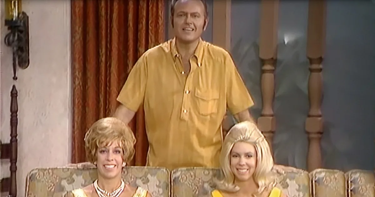 A man in a yellow shirt stands behind two women seated on a floral-patterned couch.