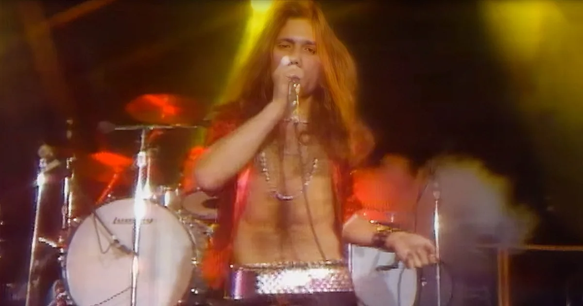 A shirtless performer with long hair sings into a microphone on stage, surrounded by musical instruments and vibrant...