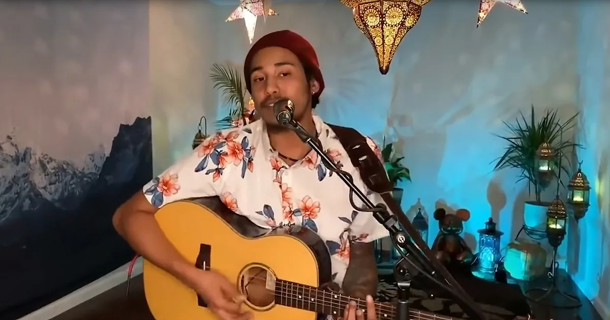 A person wearing a floral shirt and red beanie plays an acoustic guitar and sings into a microphone.