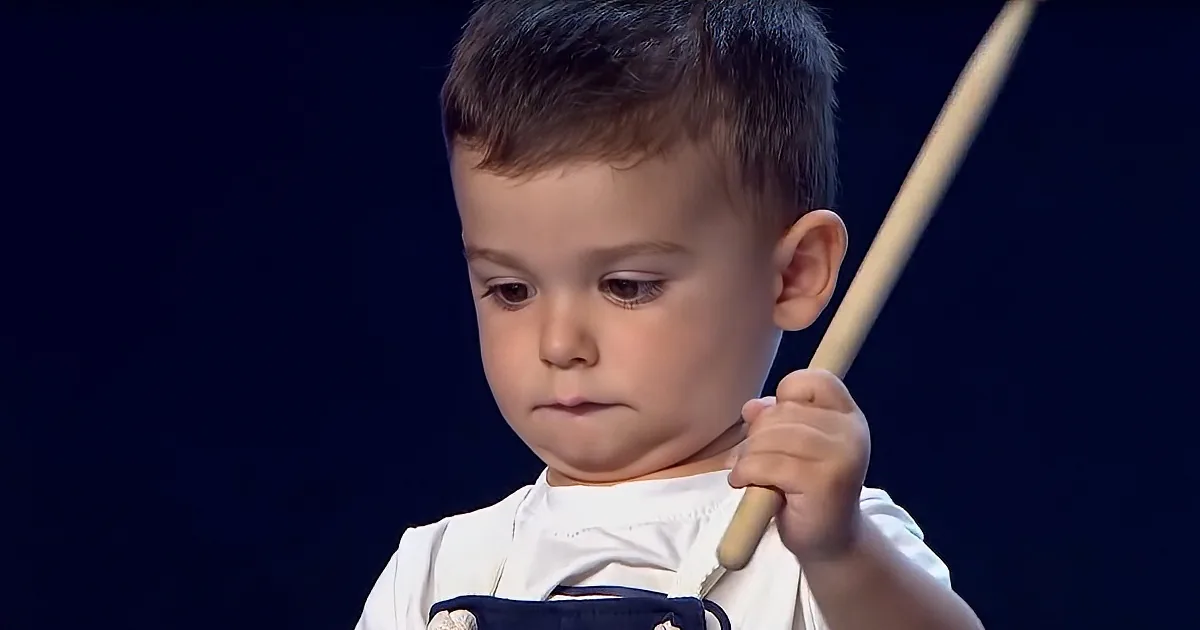 A young child in a white shirt holds a drumstick, looking down with a focused expression.