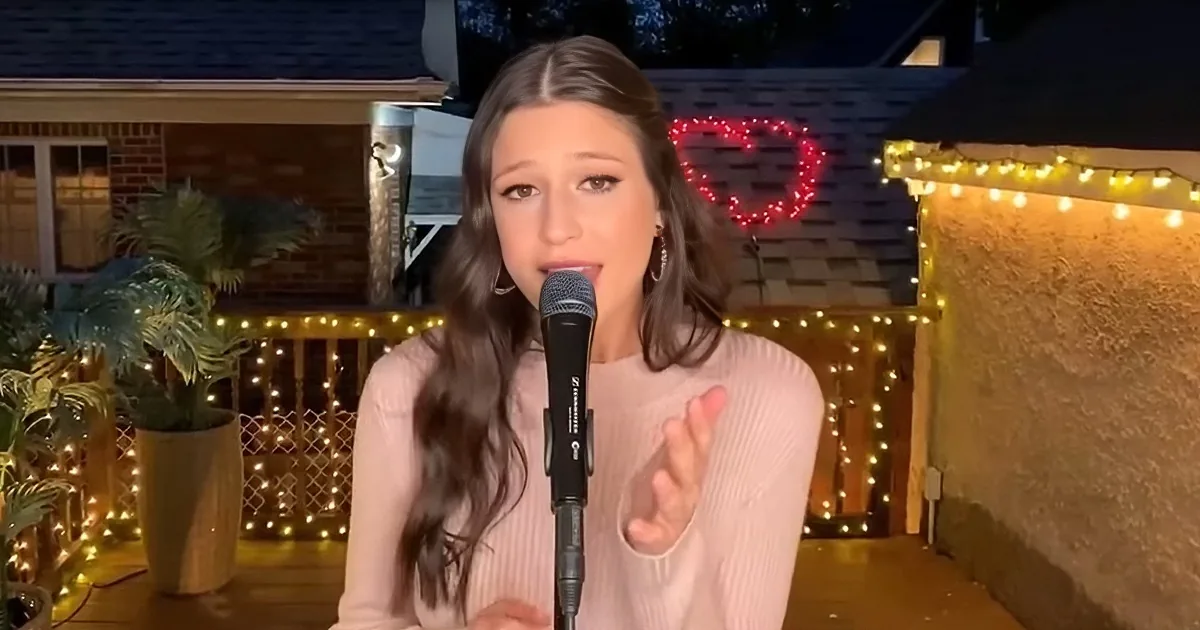 A woman with long hair speaks into a microphone, gesturing with her hand, surrounded by festive lights and plants.