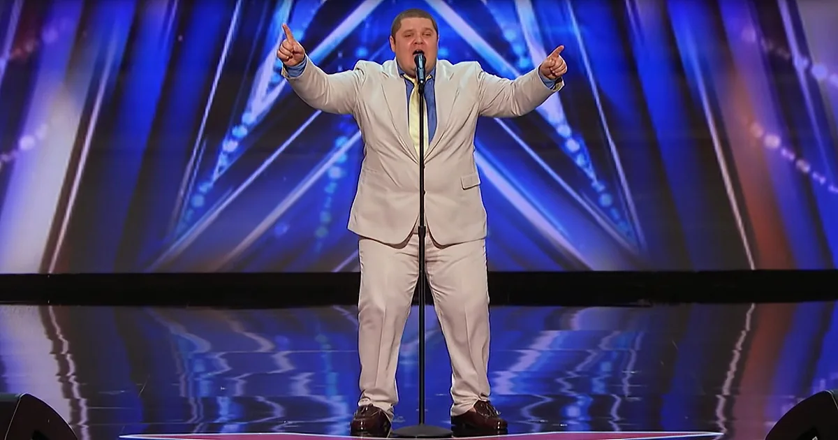 A man in a beige suit stands on stage with his arms outstretched, pointing upwards.