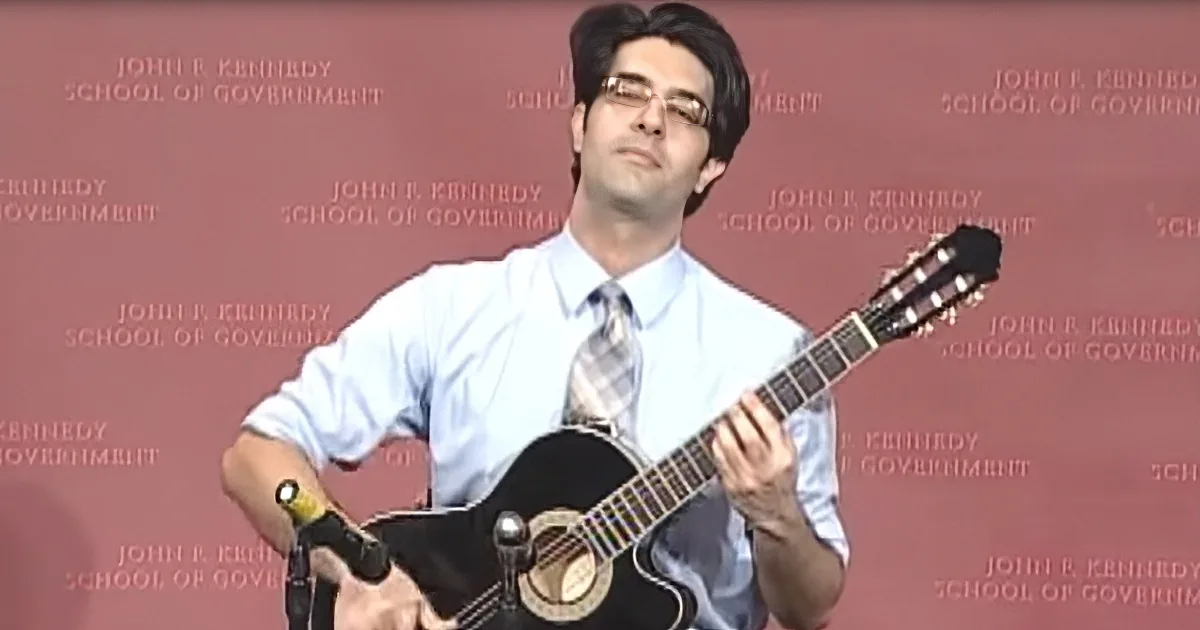 A man in a shirt and tie plays a black acoustic guitar in front of a red backdrop.