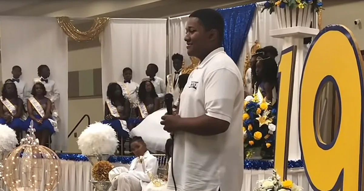 A man in white speaks into a microphone at a formal event with a large "19" decoration.