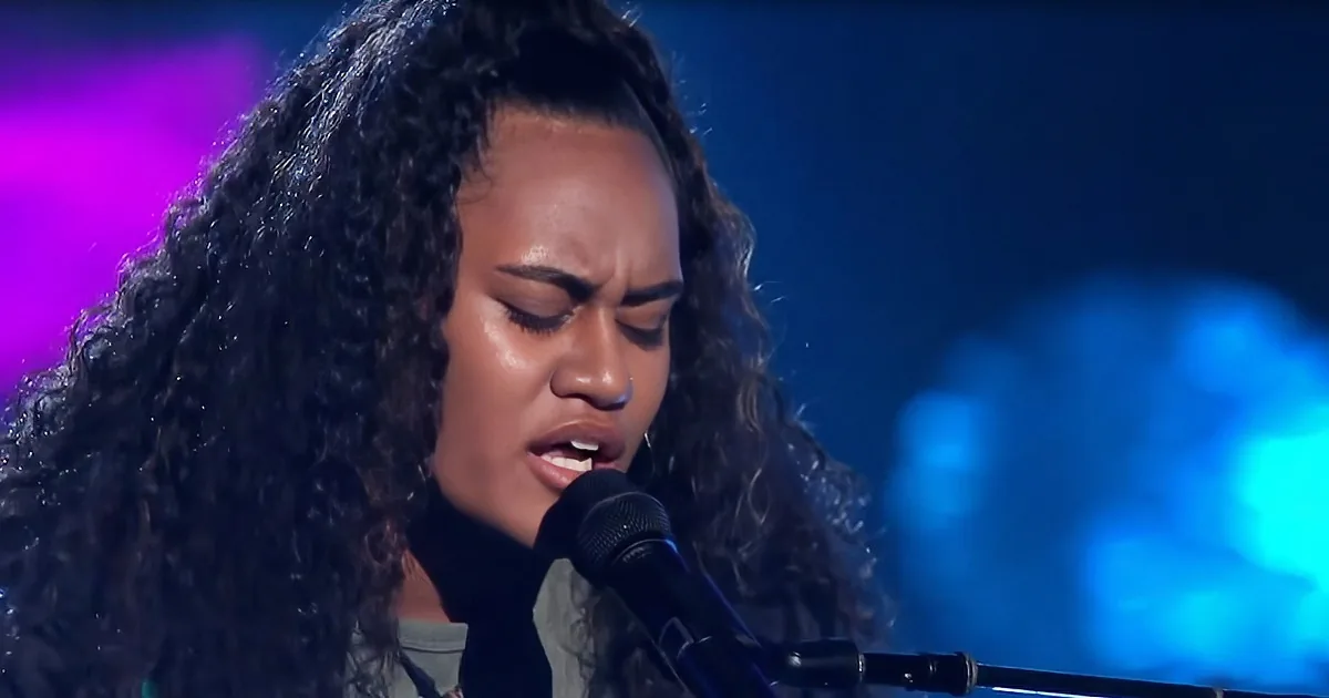 A person with curly hair sings into a microphone on a stage with blue and purple lighting.