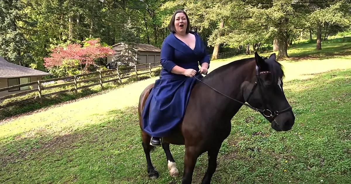 A woman in a blue dress rides a dark brown horse in a grassy, wooded area with a fence and house in the background.