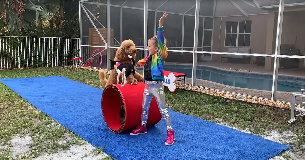 A girl in a rainbow shirt stands next to a dog in a red tube on a blue mat near a pool.