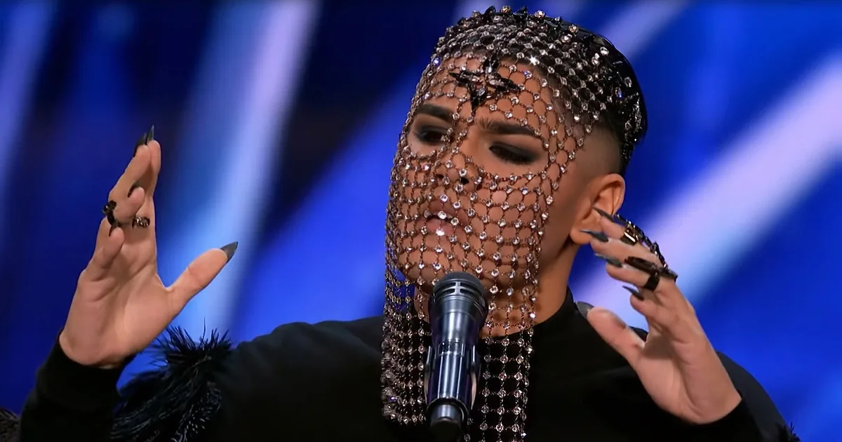 A person wearing a sparkling, beaded mask and black outfit gestures dramatically while holding a microphone.