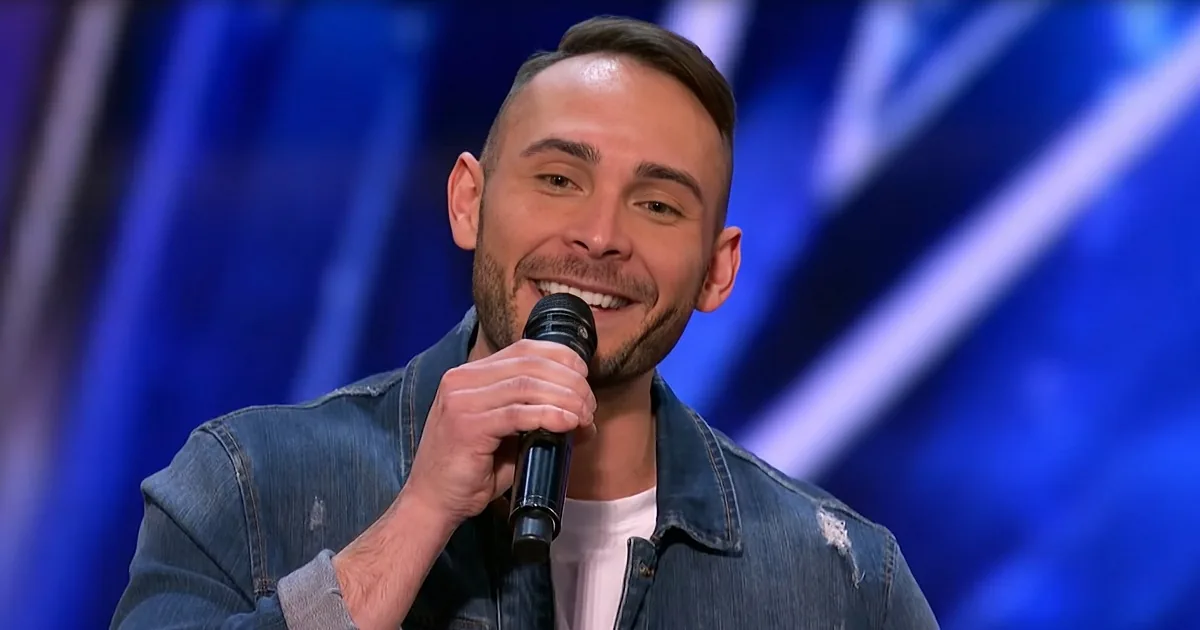 A man with a beard and short hair smiles while holding a microphone, wearing a denim jacket over a white shirt.
