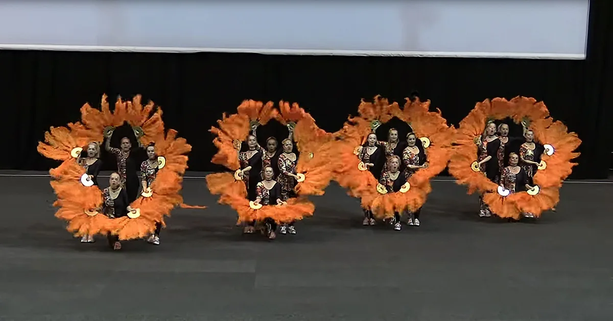 Dancers in orange fringed costumes perform a synchronized routine on stage.