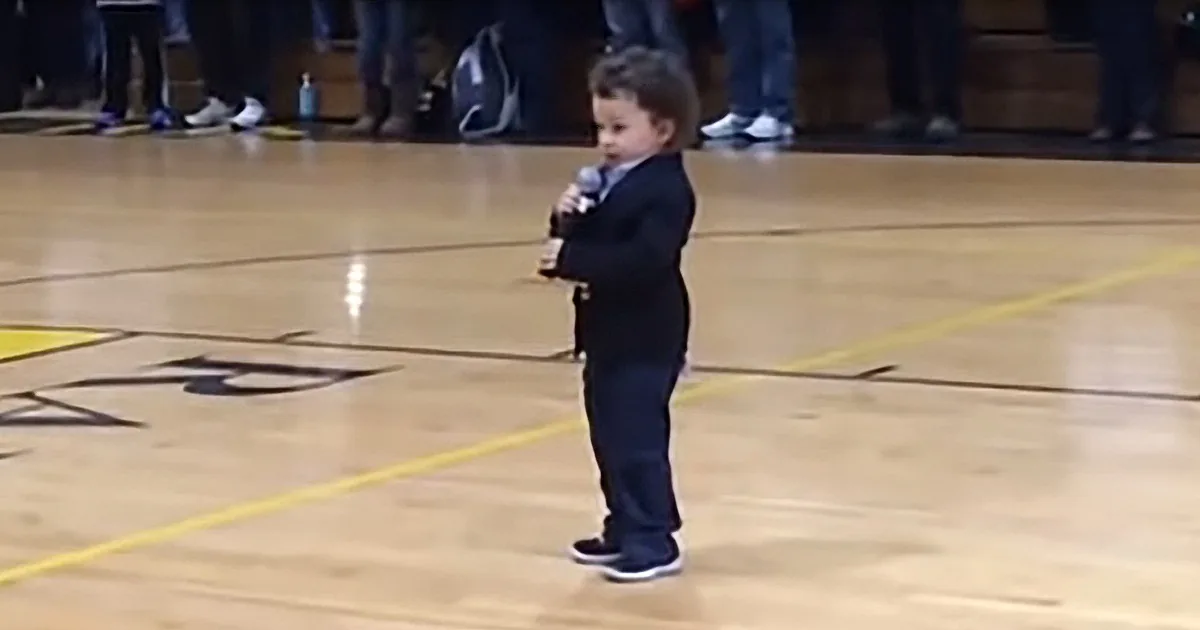 A young child in a suit stands on a gym floor holding a microphone, surrounded by spectators.
