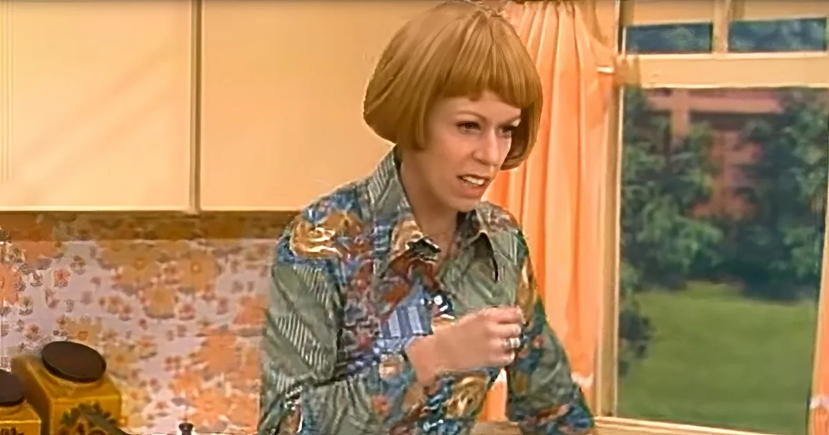 A woman with short red hair in a floral shirt stands in a kitchen with a floral backsplash and orange curtains.