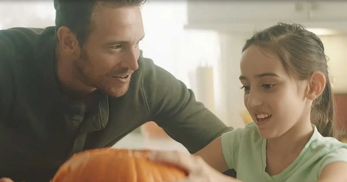 A man and a girl are smiling, carving a pumpkin together in a kitchen.